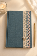 A blue textured hardcover notebook with a decorative vertical lace and ribbon trim on the right side, accented by a small rhinestone heart clasp. The notebook is styled on a light beige surface next to a gold pen, a lit candle, and soft window shadows.
